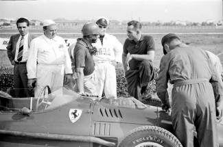 Phil Hill in sports shirt talks to Peter Collins on the grid at the 1957 Modena Grand Prix whilst portly mechanic Parenti looks on.