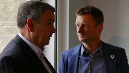 John Cleland in dark suit and white shirt on left talking to Allan McNish in blue jacket and shirt standing by a window